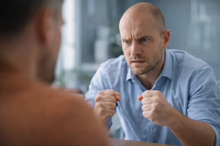 Man met gespannen blik en gebalde vuisten in een gesprek, met een onscherpe persoon op de voorgrond in een kantooromgeving.