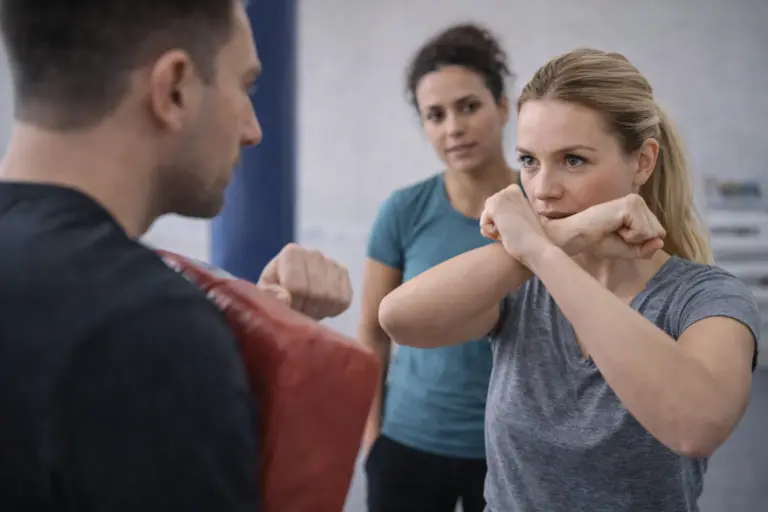Vrouw oefent weerbaarheidstechnieken tijdens een training terwijl een instructeur een stootkussen vasthoudt en een tweede deelnemer toekijkt.